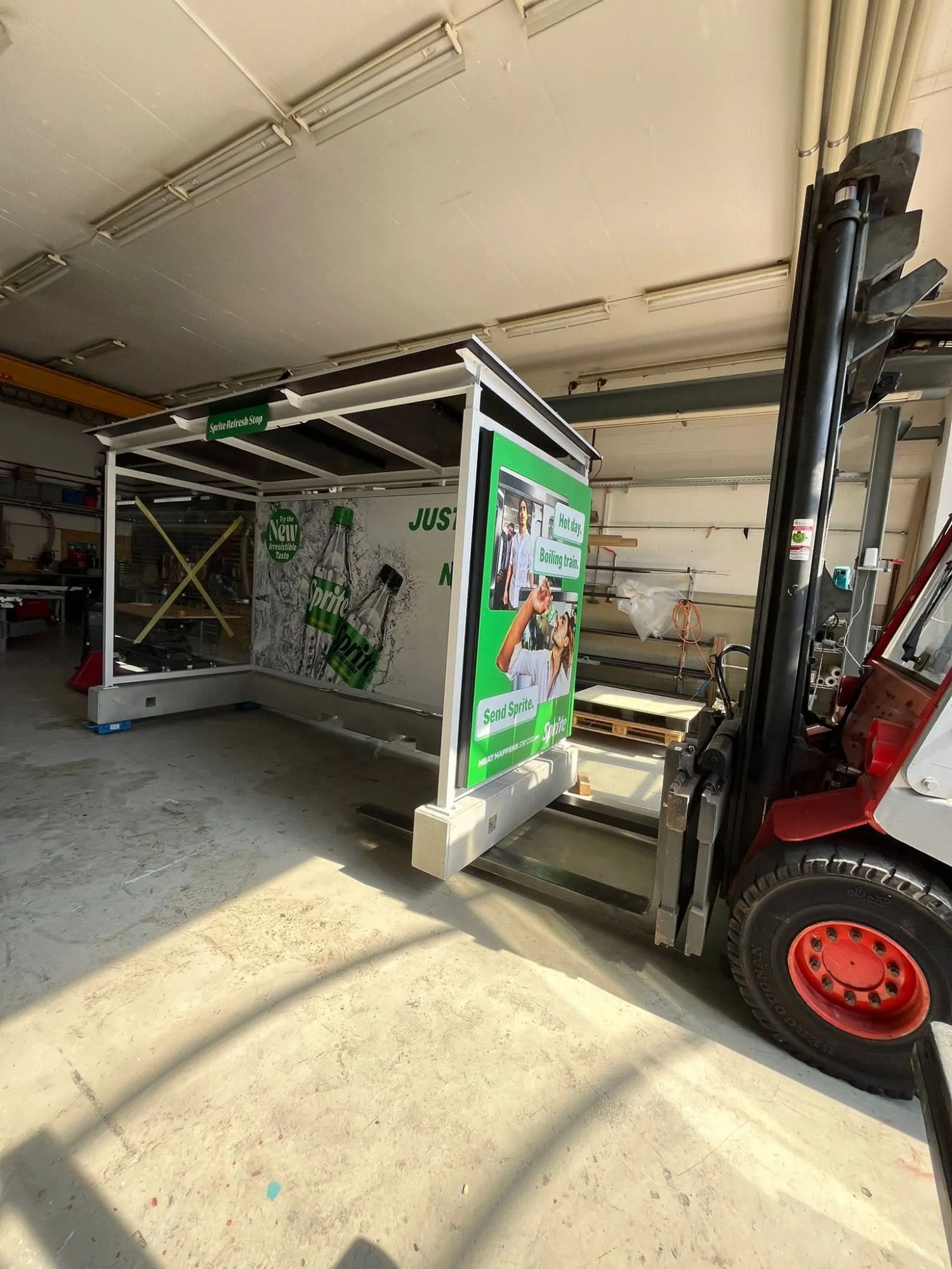 Stapler hebt die fertige Sprite-Haltestelle mit Plakat «Hot day. Boiling train. Send Sprite.» in der Werkstatt
