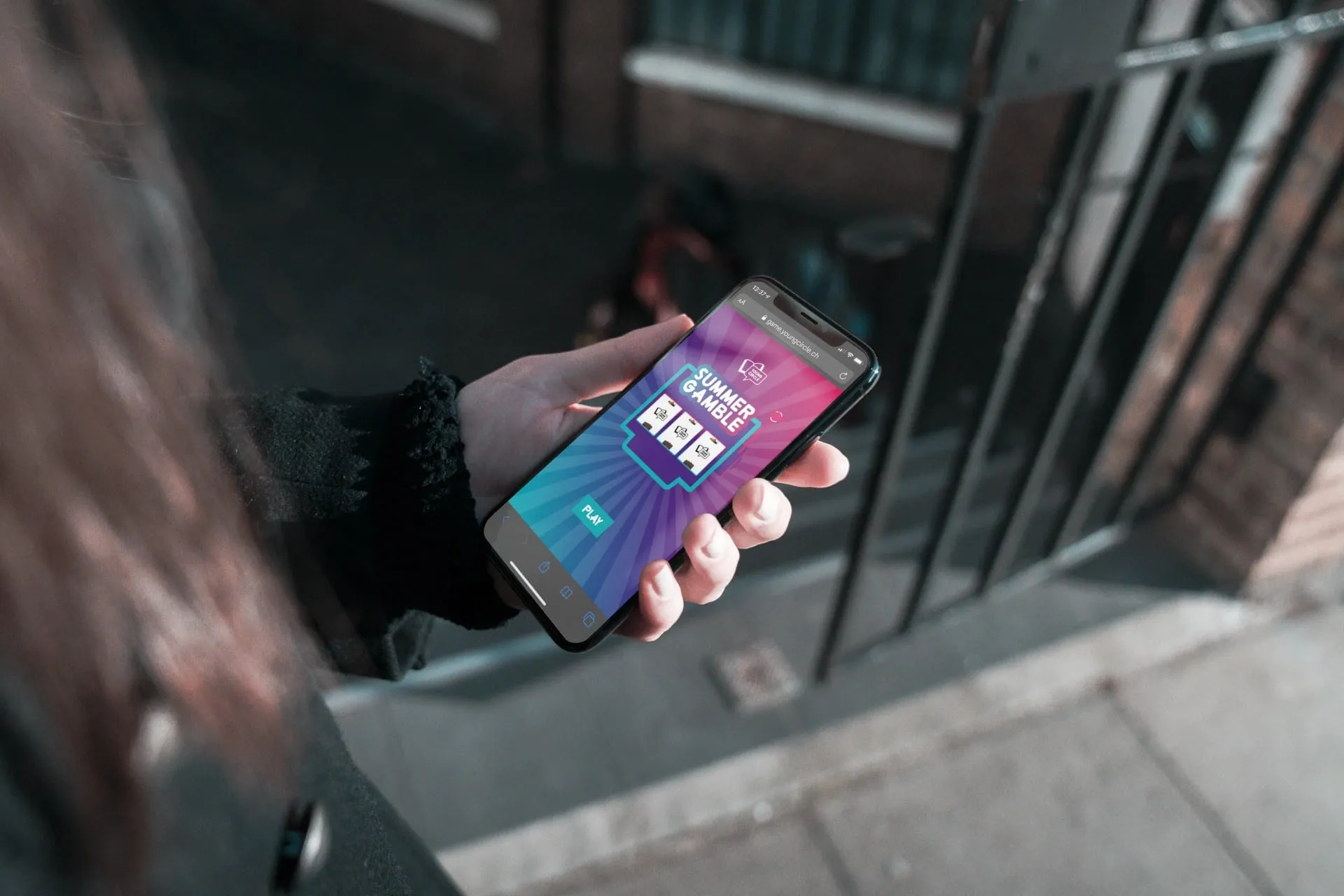 Hand holding a smartphone displaying Summer Gamble on game.youngcircle.ch, with a street in the background