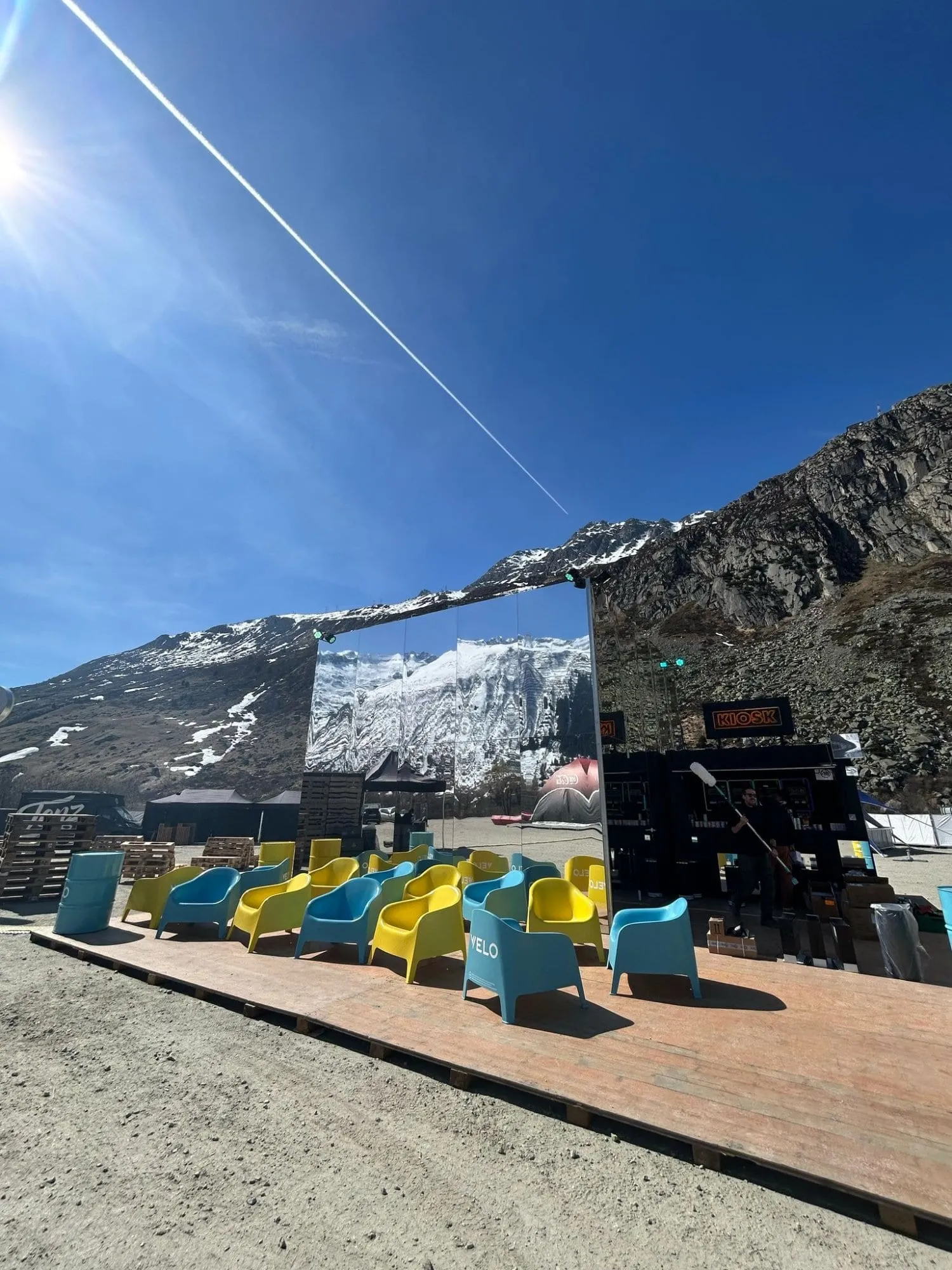 Gelbe und türkise Velo-Sessel auf dem Holzdeck vor dem Spiegel-Cube, dahinter Berge und blauer Himmel