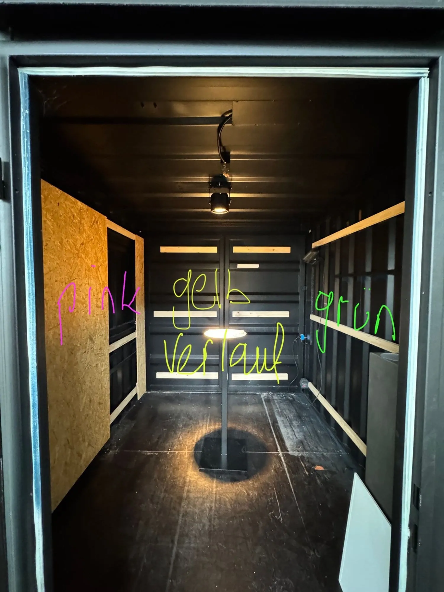 Blick in den schwarz gestrichenen Container mit OSB-Platten und Stehleuchte, mit handschriftlichen Farbnotizen «pink», «gelb Verlauf», «grün»