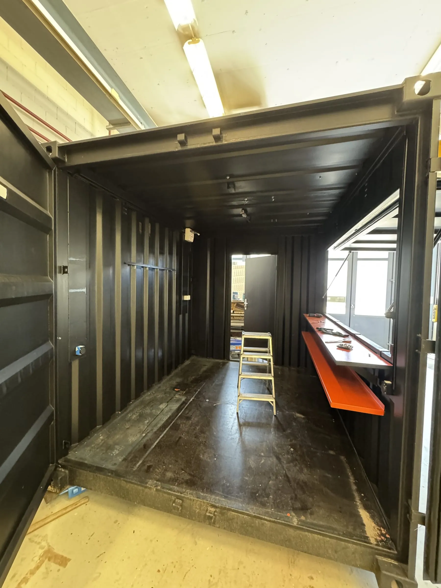 Innenraum des schwarzen Containers mit Trittleiter, orangem Thekenbrett und offener Klappe in der Werkstatt