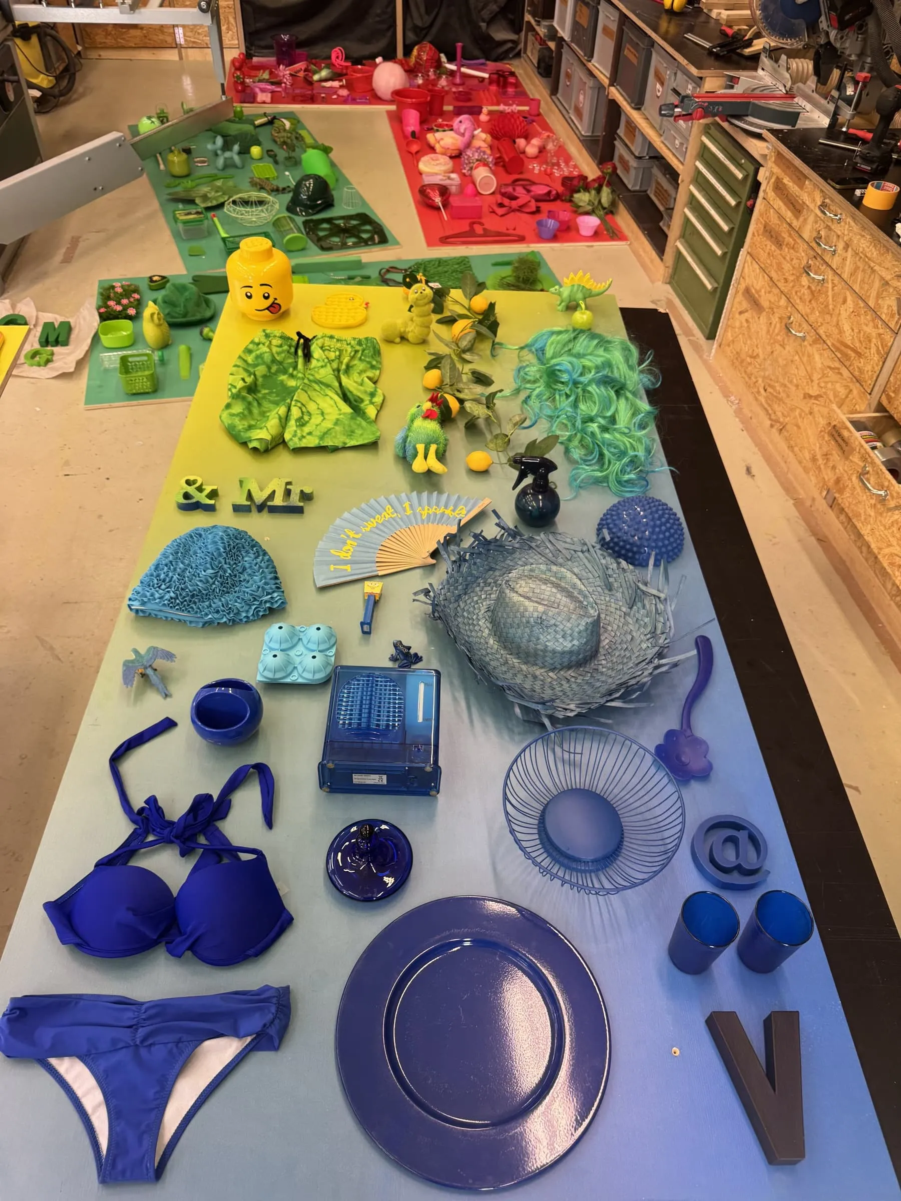 Gelb-blau verlaufende Platte mit blauen und gelben Objekten: Bikini, Strohhut, Fächer, Legokopf und Perücke