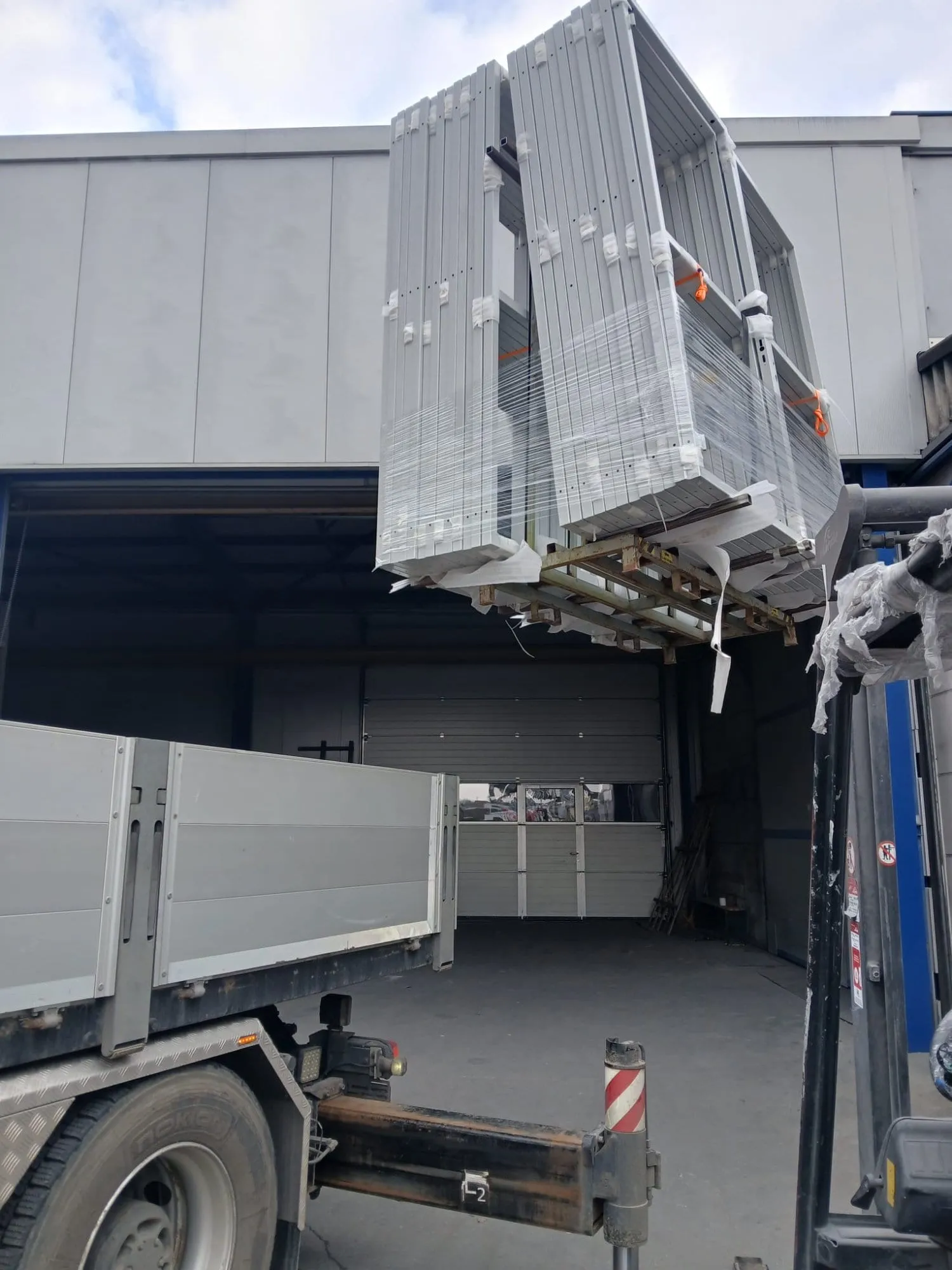 Kran hebt ein Bündel weisser Metallrahmen vom Lastwagen vor dem Werkstatttor