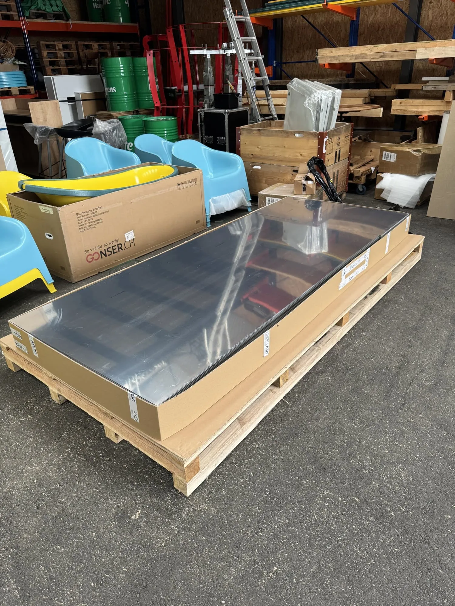 Palette mit verpackten Spiegelpanelen in der Lagerhalle, dahinter gelbe Badewanne und blaue Sessel