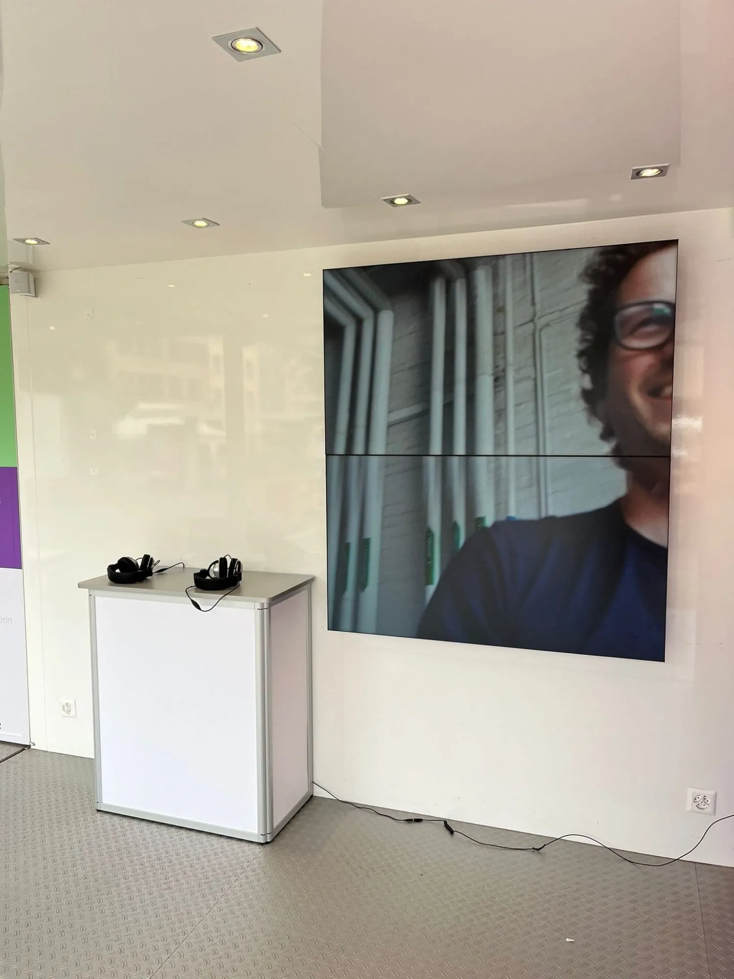 Videowall mit Grossaufnahme eines lachenden Mannes, davor weisse Theke mit zwei Kopfhörern