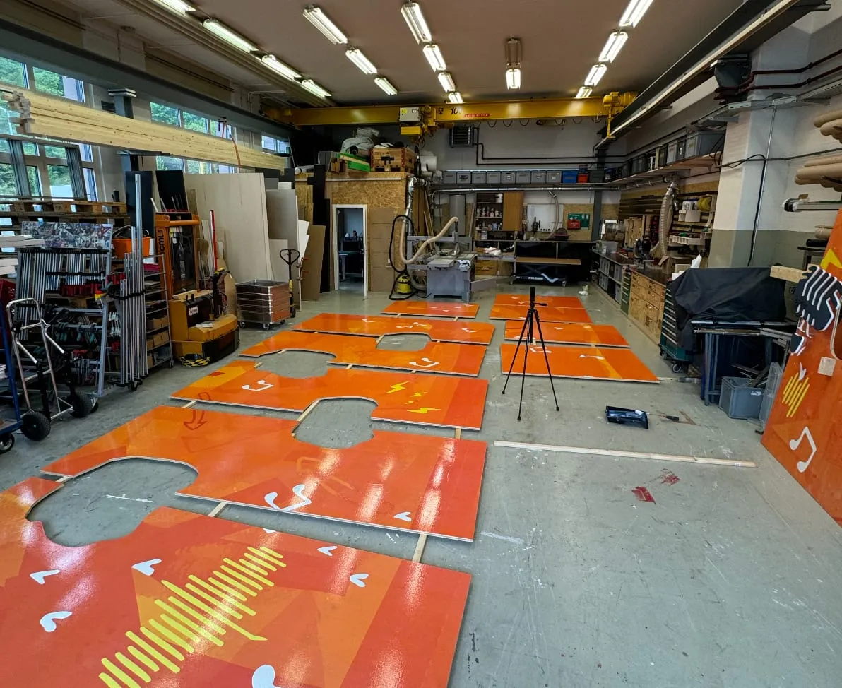Orange bedruckte Wandplatten mit Aussparungen liegen zum Zusammenbau auf dem Werkstattboden