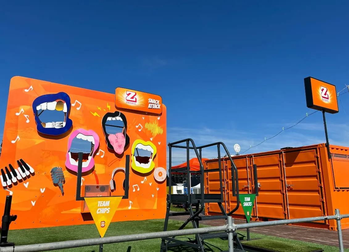 Leere orange Spielwand mit Mündern, Klaviertasten und Zweifel-Logo neben orangem Container unter blauem Himmel