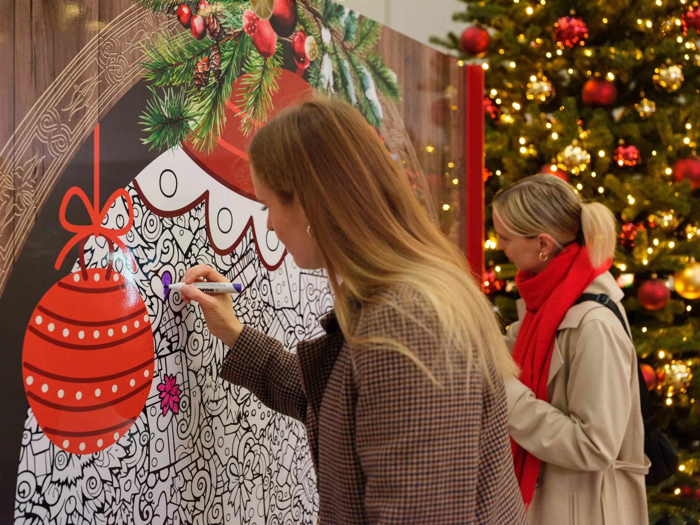 Junge Frau malt mit violettem Stift am Mandala mit roter Christbaumkugel, dahinter eine Reisende mit rotem Schal vor dem Weihnachtsbaum