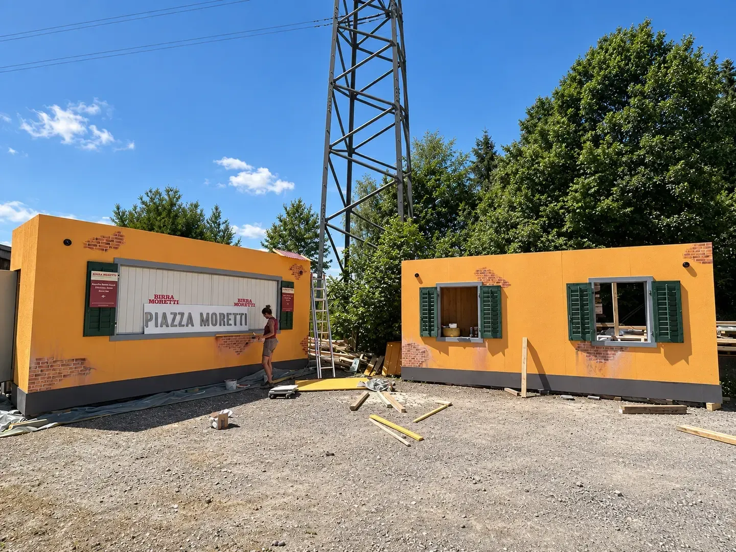 Zwei ockergelb gestrichene Container mit grünen Fensterläden im Freien vor der Werkstatt, eine Person streicht die Klappe, daneben eine Leiter