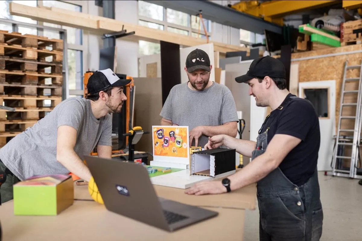 Three men in caps discussing a container model on the workbench in the workshop, a laptop next to it