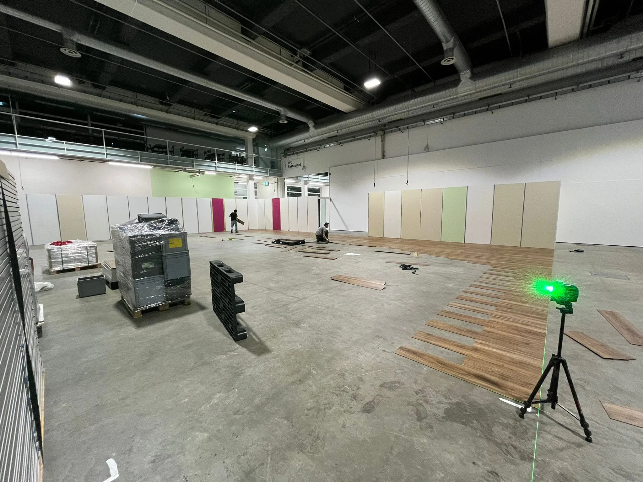 Zwei Personen verlegen Holzboden in der Messehalle, im Vordergrund ein grüner Laser auf Stativ und Paletten